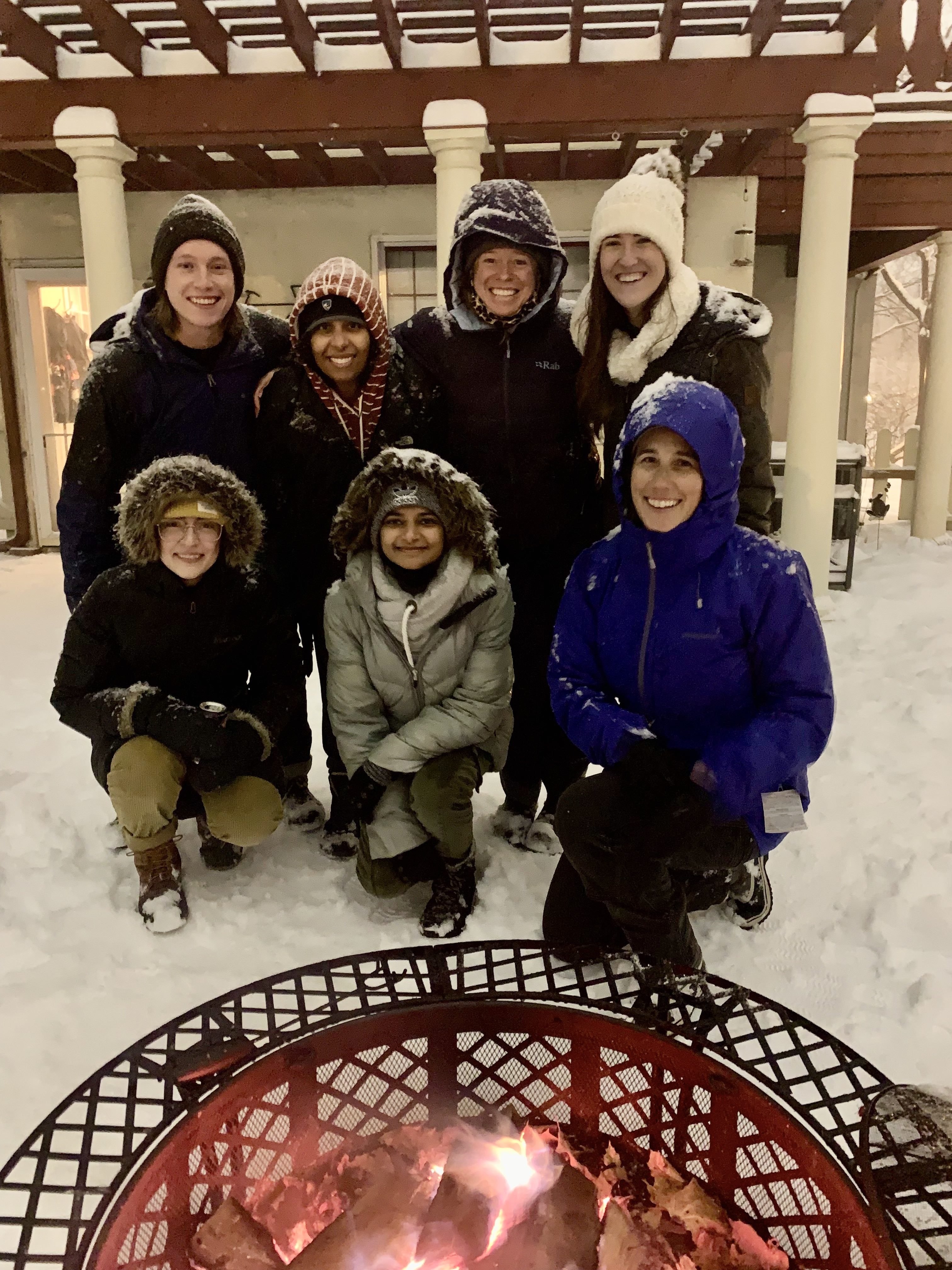 Group members and family of the Santelli Lab in front of an outdoor firepit with snow falling around them.